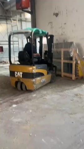 This media file depicts a forklift operator moving a pallet of items within what appears to be an industrial or warehouse setting. The forklift is yellow and black, with "CAT" branding visible on its side. A person, presumably the operator, is seated in the driver's seat, wearing dark clothing. The forklift's overhead guard is covered with a green tarp and some clear plastic sheeting.

The forklift is situated on a concrete floor that is dusty and shows signs of wear. Debris, possibly gravel or construction material, is scattered on the ground. In the background, there is a stack of items on a wooden pallet, wrapped in clear plastic. These items appear to be dark in color, possibly windows or framed materials. Another pallet with yellow items is also visible nearby.

Further back, a metal ladder or scaffolding structure is present, along with a red industrial heater mounted on the wall. The walls are concrete, with some visible damage and debris. The overall impression is a work in progress or an active industrial space. There is no indication of the time of day or weather conditions. The location cues suggest an indoor industrial environment, potentially a warehouse or construction site, consistent with the provided location context of Rozzano, Italy. The activity is straightforward material handling. No emotional cues are readily discernible.