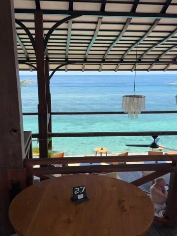 The image captures a picturesque view from an elevated, open-air restaurant or balcony, overlooking a serene tropical ocean.

In the foreground, a round wooden table with a small black sign displaying the number "27" is visible. To the right, a comfortable wicker armchair with a white cushion is positioned, suggesting a relaxed seating area. The structure is made of dark wooden beams and railings, supporting a slatted roof overhead. From the roof, two decorative, white beaded or shell-like chandeliers hang, along with a black ceiling fan, contributing to the tropical aesthetic.

Below the main viewing level, another outdoor seating area with various wooden tables and chairs is visible. A person wearing patterned shorts is partially visible in the lower right corner, near the railing of the lower deck.

The dominant feature of the scene is the ocean, which stretches out to the horizon under a bright, clear sky. The water directly below the restaurant is a vivid, translucent turquoise, hinting at a shallow, clear seabed. Further out, the water deepens into a rich, darker blue.

The scene, set in a sunny, clear weather, evokes a tranquil and inviting atmosphere, characteristic of a tropical resort or restaurant in a coastal location like Kecamatan Nusa Penida, Indonesia.
