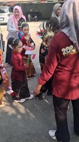 A group of children in traditional attire are walking in a line, some holding small Indonesian flags. Adults are accompanying them, with some appearing to be guiding or watching the children. The setting appears to be an outdoor area with a paved surface, possibly a street or a courtyard, with buildings and parked vehicles visible in the background. The children are dressed in various colorful outfits, some with headdresses and elaborate hairstyles. The adults are also wearing traditional clothing, including hijabs. The overall atmosphere seems to be that of a parade or a cultural event.