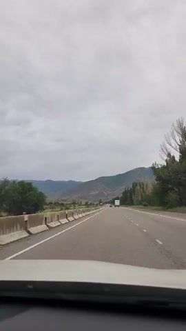 This media file depicts a scenic view from a vehicle traveling along a highway in Jil-Aryk, Kyrgyzstan. The perspective is from the driver's seat, looking forward. The road is paved and appears to be a multi-lane highway, with a concrete barrier separating it from the oncoming traffic lanes.

The surrounding landscape is characterized by rolling hills and mountains in the distance, with a cloudy sky overhead. The weather appears to be overcast, suggesting it might be a cooler day or early morning/late afternoon. Vegetation is visible on the sides of the road, including trees that appear to be somewhat sparse on the right side, possibly due to the drier climate or season. The terrain is gently sloped, typical of a valley or foothills region.

Several vehicles are visible on the highway, including what appears to be a white truck in the distance and a red bus on the left lane of the oncoming traffic. The presence of these vehicles indicates that this is a route used for transportation and commerce. There are no people clearly visible within the frame of the moving vehicle or as pedestrians.

The overall mood is one of travel and exploration, with the vast, open landscape suggesting a journey through a natural environment. The cloudy sky adds a dramatic or subdued quality to the scenery. There is no visible text or signage within the frame.