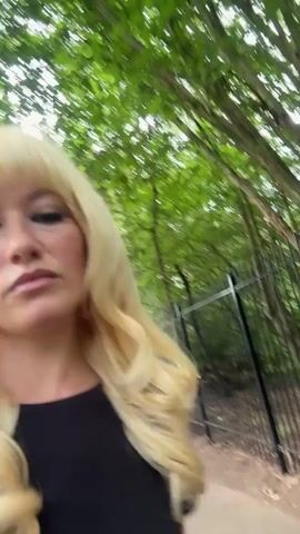 A woman with blonde hair and bangs, wearing a black top, is filmed outdoors. She has full makeup on, including dark eyeliner and lipstick. Her hair is styled in large curls. The background shows greenery, suggesting she is in a park or a wooded area, with a fence visible on the right. The camera is positioned low and close to her face, giving a slightly distorted perspective. She appears to be walking or moving, as the background is blurred. Her expression is somewhat pensive.