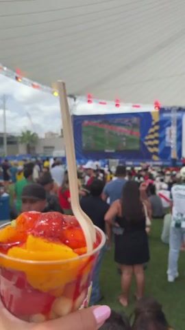 A crowd of people is gathered on a grassy area, watching a large screen that is showing a soccer match. The screen is part of a stage-like setup with banners and lights. The people are dressed casually, and some are wearing soccer jerseys. The environment appears to be outdoors, under a large white tent or awning. In the foreground, a cup with some colorful food or dessert is visible, with a spoon sticking out of it. The overall atmosphere suggests a public viewing event for a sporting match.