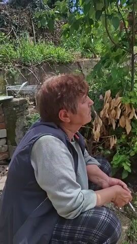 The video shows an older woman squatting in a garden, holding a cigarette. She has short, reddish-brown hair and is wearing a gray sweater, a black vest, and plaid pants. The garden is overgrown with various plants and weeds. In the background, there is a wooden structure with clotheslines attached. Beyond that, there is a glimpse of a red brick building and a blue sky with white clouds.