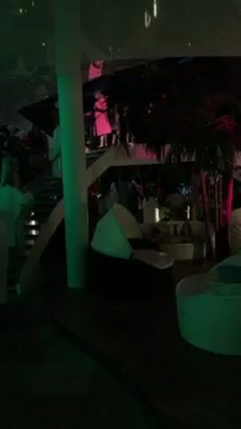 The scene takes place at night in what appears to be a luxurious, multi-level outdoor lounge area. The lighting is dim, with green and red lights adding ambiance. Several people are present, some wearing white robes, moving on stairs or standing in the upper levels. Modern white lounge furniture is visible in the foreground and background, including egg-shaped chairs and circular seating areas. Palm trees add to the outdoor aesthetic. The scene evokes a sense of relaxation and social activity.