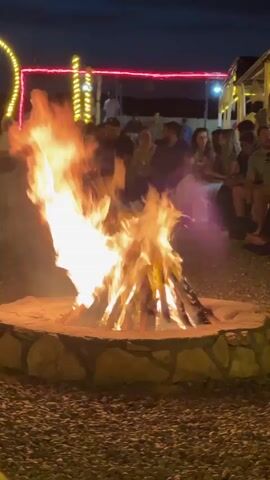 A bonfire burns in a circular stone pit, surrounded by gravel.  In the background, a group of people are seated, facing the fire. Overhead, decorative lights, including red and yellow strings, are visible.  The scene appears to be outdoors at night.