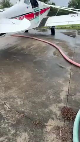 The video shows a single-engine airplane sitting on a wet concrete surface. A thick, pink hose runs along the ground near the plane's wheels. A green hose is also visible in the foreground. The plane is white with red accents. The tail number, partially obscured, appears to read "XB-TU". The weather seems overcast. The plane's engine is running.