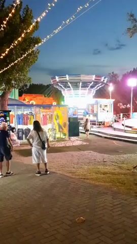 The video shows a fairground at dusk or night. String lights are strung across trees, and an amusement ride with colorful lights is in the background. Several people are visible, including two men walking on a paved pathway, and a person in a wheelchair being pushed by another person. There are booths selling merchandise, and a sign with illuminated letters reading "BALL" is visible. The overall atmosphere is festive and lively.