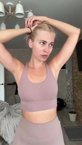 A blonde woman in a sports bra and leggings is shown from the chest up, her arms raised as she ties her hair into a ponytail. She is looking slightly to her right. The background shows a room with a light fixture, a treadmill, a bed with a blanket, a plant, and a textured wall.