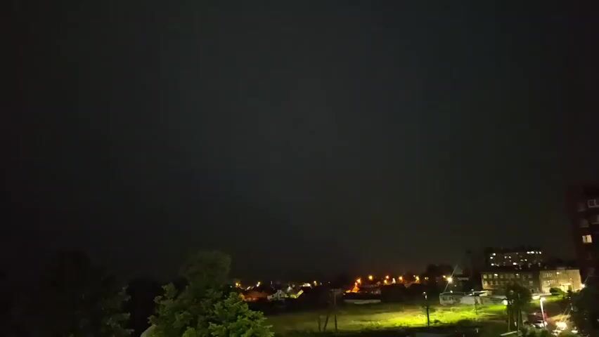 The video is a low-light recording of a nighttime scene. The sky is dark, with occasional flashes of light that illuminate the clouds and the landscape below. The landscape appears to be a rural or suburban area, with trees, houses, and streetlights visible. The flashes of light are consistent with lightning, suggesting a thunderstorm. The camera is stationary, and the framing shows a wide view of the sky and the ground. The overall mood is dramatic and somewhat ominous due to the storm.