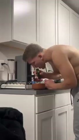 A shirtless man stands in a kitchen eating a cucumber. He is wearing grey sweatpants and has blond hair. He takes two bites of the cucumber and then smirks at the camera. The kitchen has white cabinets and a grey countertop. There is a coffee maker and a bowl on the counter. A black sweatshirt is visible on the left side of the frame.