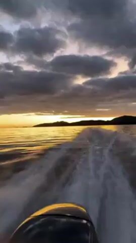 The video shows the view from the back of a moving boat on a body of water, likely the ocean. The boat is creating a wake of white water behind it. In the distance, there are dark landmasses silhouetted against a golden sunset. The sky is filled with dark, greyish clouds, with the sun's rays breaking through to illuminate the water. The boat's black motor is visible in the foreground.