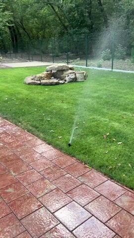 A sprinkler sprays water onto a green lawn in front of a rock formation that appears to be a decorative waterfall. A tiled patio is visible in the foreground. Trees and a fence are in the background.