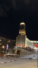 Woman photographs illuminated clock tower with red flags at night