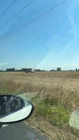 The video shows a blue tractor pulling a large tank through a dry, harvested field under a clear blue sky. The field appears to be agricultural land, with remnants of crops visible. In the distance, there are trees and buildings, suggesting a rural setting. The camera seems to be filming from inside a moving vehicle, as indicated by the side mirror visible in the lower left corner of the frame. The tractor is the primary focus, moving across the landscape.