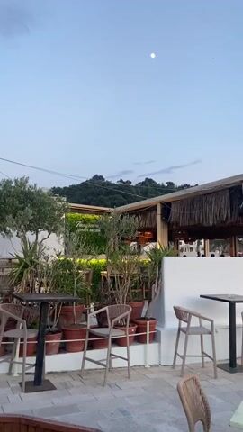 The video shows an outdoor patio with tables and chairs, part of a restaurant overlooking the sea. The setting is dusk, with the sky transitioning from light blue to shades of pink and orange on the horizon. The water is calm, and a small, rocky island is visible in the distance. People are seated at some of the tables, enjoying the view. The patio is decorated with plants and features a covered area with a thatched roof.