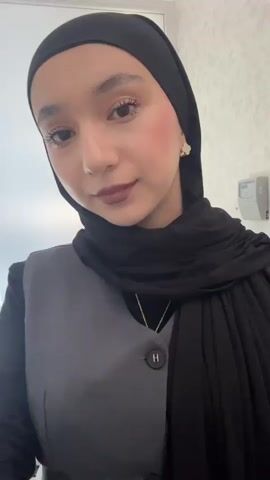 A woman with fair skin and brown eyes is shown from the chest up. She is wearing a black hijab, a dark grey vest with one button, and a black shirt underneath. She is also wearing a gold necklace with a small pendant. She is standing in a room with white walls, and a few items are visible in the background, including what appears to be a light switch and some items on a shelf. The woman is looking directly at the camera with a slight smile.
