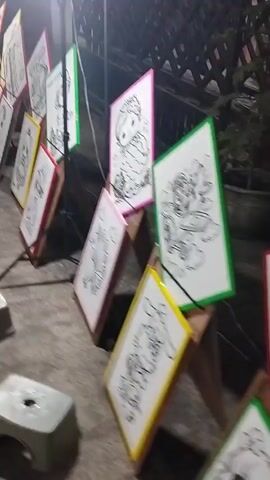 The video shows a row of coloring pages displayed on easels. The coloring pages are framed and have colorful borders. Some of the coloring pages are of cartoon characters. There are several small gray stools in front of the easels. The scene appears to be outdoors, possibly at a market or fair, with a dark background suggesting it may be nighttime or an indoor setting with dim lighting. The camera is moving, giving a slightly blurred view of the scene.