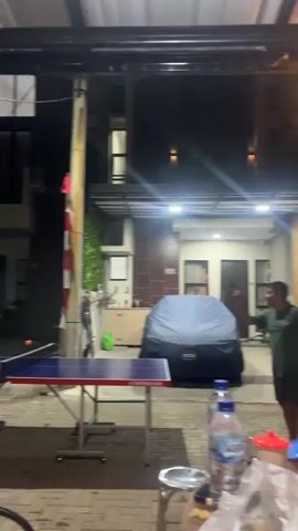 A low-angle, nighttime outdoor shot shows a portable ping pong table set up on a paved surface under a covered patio. Two men are playing ping pong. The first man, shirtless and wearing black pants, hits a ball across the table. The second man, wearing a dark green t-shirt and dark pants, smiles as he prepares to hit the ball back. A bicycle is visible to the left of the frame, and a car covered with a blue tarp is in the background to the right.
