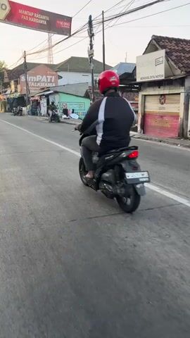 A person wearing a red helmet and a black and white jacket rides a black scooter down a paved road. The scooter has a white line on its rear. The road has a white line marking the lane. On the left side of the road, there are several buildings and a sign that reads "simPATI TALKMANIA." On the right side of the road, there are more buildings and a sign that reads "smartfren." The sky is a pale orange, suggesting it is either sunrise or sunset. The environment appears to be a town or city street.