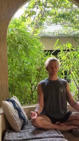 A medium shot shows a man meditating outdoors on a cushioned surface, with lush green foliage in the background. The man is seated in a cross-legged position, eyes closed, and hands resting on his knees in a meditative pose. He has light skin, his hair is pulled back in a small bun, and he has tattoos on his arms. He is wearing a gray tank top and dark shorts. To his left is a decorative pillow with a blue and white pattern. A small wooden table is visible in the right foreground, holding a glass of a red liquid. The setting appears to be a covered patio or alcove, with bamboo plants and other greenery creating a tranquil atmosphere. The camera is static.