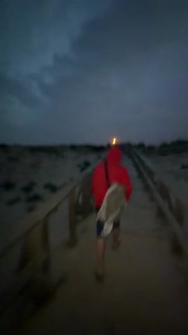The video depicts a sandy beach landscape at dusk, under a cloudy sky. The camera seems to be moving along a wooden path towards the ocean, as evidenced by the blurred wooden railings in the foreground. The sea is visible in the distance, with small waves breaking. There is a person in the frame towards the right in a red hoodie. In the latter half of the video, a hazy moon is visible through the clouds. The scene is somewhat dark, with limited visibility due to the low light.