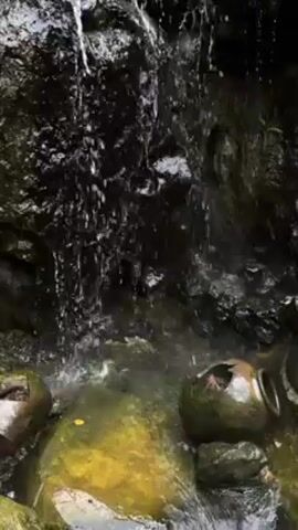 The video shows water trickling down a dark, moss-covered rocky wall into a small pool.  The pool is partially covered with green-tinged stones and contains several broken, dark-colored ceramic pots.  The focus is on the falling water and its effect on the pool.