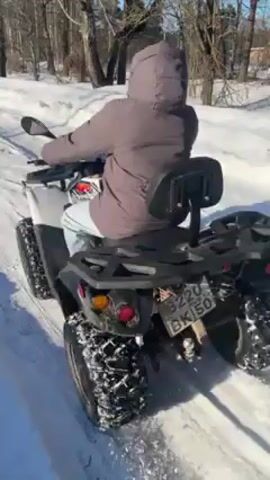 Here is a concise description of the video:
A person wearing a winter coat and hood rides a white ATV on a snow-covered path. The ATV has a black rear rack, and its license plate is visible. The path is flanked by snow and trees on either side, under a bright blue sky. The ATV drives away from the camera.