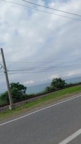 The video shows a view from a vehicle traveling alongside a railway track and the sea. The sky is cloudy. There are power lines overhead, supported by poles. The railway tracks run parallel to the road, with a grassy embankment between them. In the distance, the sea stretches out to the horizon. The video captures a section of the railway line, including a small bridge-like structure over the tracks. No people are visible in the video. The environment appears to be coastal, with the sea on one side and land with vegetation on the other.