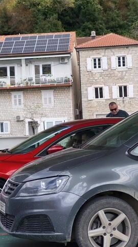 A man with sunglasses is standing next to a gray car parked next to a red car. Another person is sitting inside the red car. The man is looking down at the car next to the red car. In the background are two buildings with white stone facades and trees above them. The building on the left has solar panels on the roof.