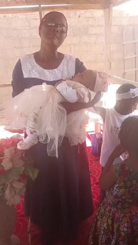 A woman stands holding a baby in a white dress and a pink headband. The woman is wearing glasses, a red head covering, a black and white top and a black skirt. Three children stand next to her looking at the baby. One child is wearing a white top and a white headband, and the other child is wearing a multicolored floral dress. The third child's head is only partially visible. The woman appears to be speaking. There is a floral arrangement to the left of the woman. The scene appears to be indoors under a roof. The walls are concrete blocks. There is a red rug on the floor.
