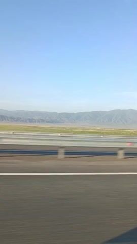 A low-angle shot captures a highway guardrail and the road as a vehicle moves forward. The guardrail is made of silver metal with reflective red markers. Beyond the guardrail, a vast, flat plain stretches towards a range of distant, hazy mountains under a clear blue sky. The terrain appears arid and sparsely vegetated. The focus is on the movement and the expansive landscape.