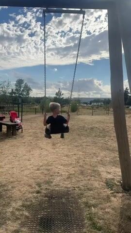 A young boy with blond hair, wearing a dark blue t-shirt and shorts, is swinging on a swing set. The swing has a black seat and metal chains. The boy is facing away from the camera and appears to be pushing himself to swing higher. The background shows a dry, grassy field with some trees and a cloudy sky. There is a wooden picnic table with a red backpack on it to the left of the frame. The sun is shining brightly, creating lens flare in the upper right corner of the video. The swing set is made of thick wooden posts.