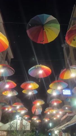 The video captures a vibrant night scene on a narrow urban street in Karditsa, Greece. The sky is dark, indicating it is nighttime. Overhead, a multitude of rainbow-colored umbrellas are strung across the street, forming an illuminated canopy. Many of these decorative umbrellas have bright lights positioned beneath them, casting a warm glow and creating a festive atmosphere.

On either side of the street, multi-story buildings with various shopfronts and awnings are visible. Some green foliage, possibly from planters or small trees, lines the street at ground level. Bright streetlights also contribute to the overall illumination of the scene.

Towards the end of the video, a person can be briefly seen walking along the right side of the street. Several signs with Greek text are visible on the buildings. One blue sign, though partially obscured by lighting, appears to include "ALOMARAS & PARTALIDIS" and additional Greek text. Below it, a yellow sign clearly reads "ΕΚΔΟΣΕΙΣ ΓΡΑΨΙΜΟ" (Publications Writing). The camera maintains a relatively steady, slightly upward-tilted view, showcasing the colorful umbrella display.