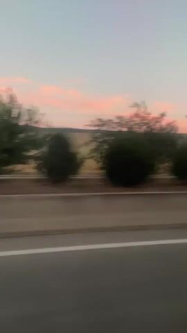 The video shows a blurred view from a moving vehicle, likely a car, traveling on a highway. The scene is captured at dusk, with a soft pink and orange sky visible in the background. There are trees and rolling hills in the distance. The highway itself is visible in the foreground, with a white lane marking. The motion blur indicates that the vehicle is in motion, creating a sense of travel.