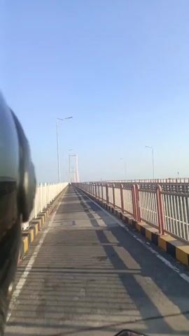 The video shows a first-person perspective of someone riding a motorcycle across a suspension bridge. The bridge has a paved roadway with distinct lanes marked by yellow and black stripes along the edges. White and red railings line both sides of the bridge, and streetlights are visible at intervals. The sky is clear and blue, indicating daytime. In the distance, the bridge's main towers are visible against the horizon. The rider's helmet and part of the motorcycle are visible in the foreground. The motorcycle is moving forward on the bridge, casting long shadows on the roadway due to the sunlight.