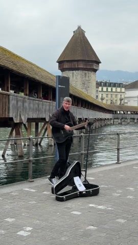 A man with short gray hair plays a black acoustic guitar and sings near a river. He is dressed in black and stands near an open guitar case and a music stand. Behind him is a wooden bridge with a tower at one end, typical of Lucerne, Switzerland. The river runs alongside the bridge, and buildings are visible in the background under a cloudy sky.
