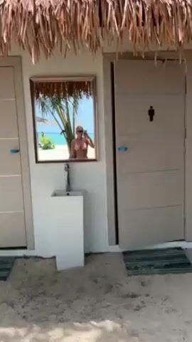 The scene is a public restroom on a tropical beach. The restroom is white, with two gray doors, one labeled with a male symbol. A small sink is mounted on the wall between the doors, and a mirror hangs above the sink. The mirror reflects a woman in a bikini, who is taking a selfie. The reflection also shows a white sand beach, blue water, and palm trees. The restroom has a thatched roof.