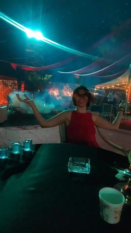 A woman with short hair sits at an outdoor table covered in a dark green cloth. She is wearing a red sleeveless top and has her arms outstretched with palms up, a smile on her face. On the table in front of her are several small, teal-colored glasses, an ashtray, and a paper cup. The background is a dimly lit outdoor event space, decorated with hanging red and white fabric, string lights, and what appears to be a smoke effect. A bright light is positioned above, creating a lens flare. Other people and seating areas are visible in the background.