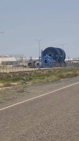 The video shows several large, blue spools of cable stacked outdoors. The spools are of varying sizes, with the largest being significantly taller than a person. They are arranged in a somewhat haphazard manner. Behind the spools, there is a fence and some buildings that appear to be industrial or storage facilities. The sky is clear and blue, suggesting a sunny day. The ground in the foreground is a paved road, and there is vegetation on the side of the road. The video appears to have been recorded from a moving vehicle. No people are visible in the video.