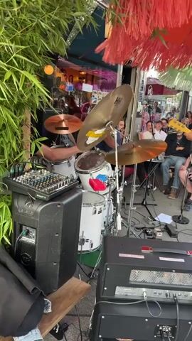 A drummer, positioned behind a white drum kit with a silver cymbal, plays the drums in an outdoor setting. The drummer, wearing a dark shirt, is visible from the waist up, with their arms in motion as they strike the drums and cymbals. To the left of the drum kit is a black speaker with a silver grille and a green mixer with numerous knobs and sliders. A wooden plank is placed in front of the speaker. In the background, behind the drummer and drum kit, a band appears to be performing. People are seated in chairs, facing the band. Some are looking at the performers, while others are engaged in conversation. The environment suggests an outdoor cafe or restaurant with tables and chairs set up. A bamboo plant with green leaves is visible in the foreground on the left side of the frame. A red fringed canopy hangs overhead, partially obscuring the view of the background. The lighting is bright, indicating it is daytime.