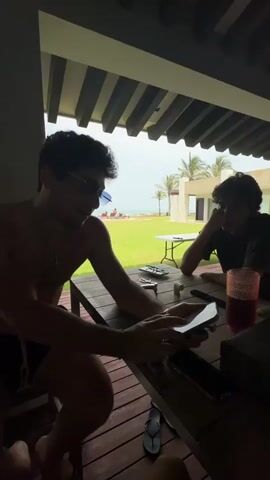 The video captures three young adult men relaxing on a covered outdoor patio of a residence in Mata de Uva, Mexico. They are gathered around a wooden table.

Initially, two men are seated; one is focused on his smartphone, while the other observes. The camera then pans, revealing a third man standing beside the table. The man who was on his phone looks up and smiles at the camera, while another man seated opposite him also looks towards the camera. The scene suggests a casual social gathering. One man is later shown eating from a plate of green-colored food, possibly scrambled eggs or chilaquiles.

On the table, there are smartphones, a dark decorative case, red plastic cups, a glass with a dark red drink, a water bottle labeled "WONDER," a remote control, and a plate of food. A pair of black flip-flops is visible on the floor.

The setting is bright daytime, likely under an overcast or cloudy sky, as natural light illuminates the scene without harsh shadows. Beyond the patio, a spacious, well-maintained green lawn leads to several palm trees and the clear blue ocean in the distance. Large glass doors separate the patio from an elegant interior living space. The overall atmosphere is relaxed and casual.