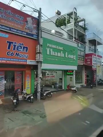 The video shows a street scene with several storefronts. On the left, there is a green and white sign for a dental clinic named "Thanh Long". Next to it, there is a red and white sign for a clinic called "Chi Mi" which offers cosmetic dentistry. Further down the street, there is a sign for a clinic named "Nguyen Van Chin" specializing in dental and cosmetic procedures. To the right, a white taxi is parked on the street. Beyond the taxi, more storefronts are visible, including "Shop Linh San 18" and "Lucy", which appears to be a beauty salon. There are several motorbikes parked along the sidewalk in front of the shops. A few people are visible walking on the sidewalk. The sky is cloudy.