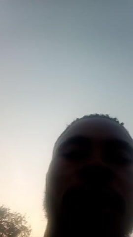 The video shows a Black man speaking directly to the camera. He has a short beard and mustache, and he is wearing a brown t-shirt. He has a bag slung over his shoulder.  The background is a street scene with buildings and a few cars. The light is bright, and the man is looking into the light. He is speaking enthusiastically.