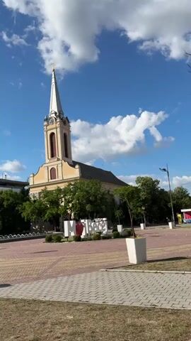 The video displays an outdoor scene featuring a large, light-colored church with a tall, white steeple topped with a golden cross. The church has a brown roof and rectangular window or decorative elements below the steeple. In front of the church is a paved public square with a patterned surface. Several green trees are visible around the square and partially obscuring the church. To the right of the church, a white sign with a red heart reads "I LOVE BEČEJ". The sky is blue with scattered white clouds. The foreground shows a patch of dry grass and white planter boxes on the edge of the paved area. The scene is brightly lit by sunlight.