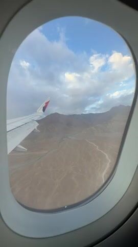 The video shows a view from an airplane window as it flies over a mountainous, arid landscape. The wing of the airplane is visible in the foreground on the left side of the frame. The wing is white with a logo that includes red, white, and purple colors. The landscape below consists of brown, rocky mountains and a dry riverbed or ravine winding through the valley. The sky is partly cloudy with patches of blue visible. The camera angle and movement suggest that the airplane is either ascending or descending, or maneuvering through the mountainous terrain. The lighting indicates it might be early morning or late afternoon. There are no people visible in the video.