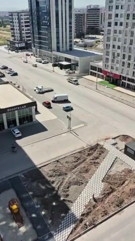 The video is an aerial shot of a street with several buildings and a playground. On the left side of the street, there is a building with the words "SPORTS LAB" visible on its facade. In front of this building, a white car is parked. Further down the street, there are several parked cars and moving vehicles, including a white truck and a white van. On the right side of the street, there are tall apartment buildings. In the foreground, there is a playground area with various pieces of equipment, including what appears to be a slide and several wooden structures that look like agility training obstacles for dogs. The ground in the playground area is covered with gravel and some dirt, indicating it might be under construction or recently completed. The overall environment appears to be an urban setting with a mix of commercial and residential buildings.