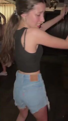 A medium shot captures a young woman dancing in what appears to be a dimly lit indoor space. She has long brown hair tied back in a ponytail with a black and white scrunchie. She is wearing a black tank top and light blue denim shorts with a frayed hem. Her back is to the camera as she moves, her hair swishing from side to side. Her right arm is raised above her head, and her left arm is extended outwards. She glances over her shoulder, smiling. In the background, another person is visible on the left, also dancing, and various objects including a coat rack and what looks like a bar or counter are present. The floor is dark wood.