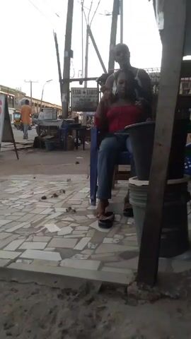 A young woman in a red top and jeans sits in a blue plastic chair on a tiled sidewalk, having her hair braided by another young woman in a white top. The braider stands behind the seated woman, working on her hair. The scene is outdoors, on a street with other people and objects visible in the background, including a sign for "STAR LODGE," power poles, and miscellaneous buckets and items. The overall lighting suggests it might be early morning or late afternoon.