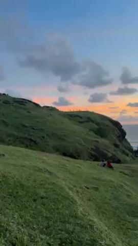 The video shows a lush green hillside under a sky painted with the colors of sunset. The horizon is a mix of orange, pink and purple hues.  Several people are standing on the crest of the hill, silhouetted against the colorful sky. At the bottom of the hill, nearer to the camera, are two individuals sitting on the grass. The environment appears tranquil and scenic.