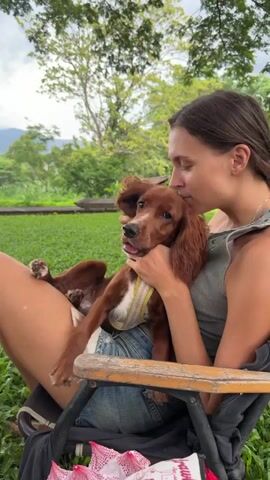 A young woman with brown hair tied back is sitting in a chair outdoors, holding a small brown dog in her lap. The dog is wearing a pink and white harness. The woman is leaning her head towards the dog, appearing to be kissing or nuzzling it. The dog's mouth is open, and it looks happy. They are in a grassy area with trees and foliage in the background. The lighting suggests it is daytime.